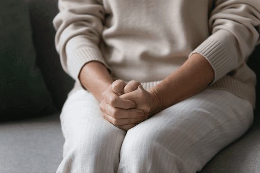 A woman sitting on a couch, her hands clasped tightly together, displaying signs of anxiety.