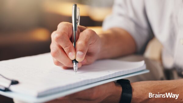 Close-up of a person writing notes on a clipboard, with the BrainsWay logo.