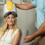 Woman sits in a treatment chair wearing a BrainsWay TMS helmet while a clinician adjusts the device.