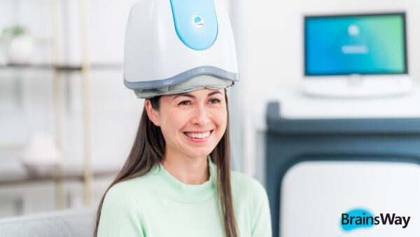 Smiling woman seated in a clinic wearing a white BrainsWay Deep TMS helmet, with a treatment monitor behind her.