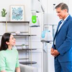A smiling woman sits in a gray armchair, talking with a man in a blue suit in a treatment room with BrainsWay Deep TMS helmet devices.