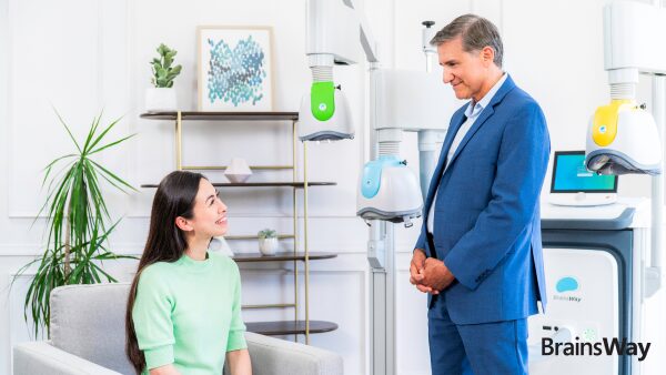 A smiling woman sits in a gray armchair, talking with a man in a blue suit in a treatment room with BrainsWay Deep TMS helmet devices.